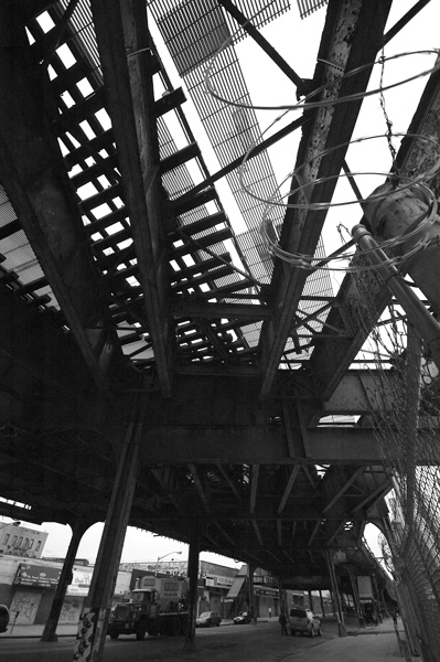 An elevated train track casts darkness on a street, what with its myriad rails, supports, and work platforms.