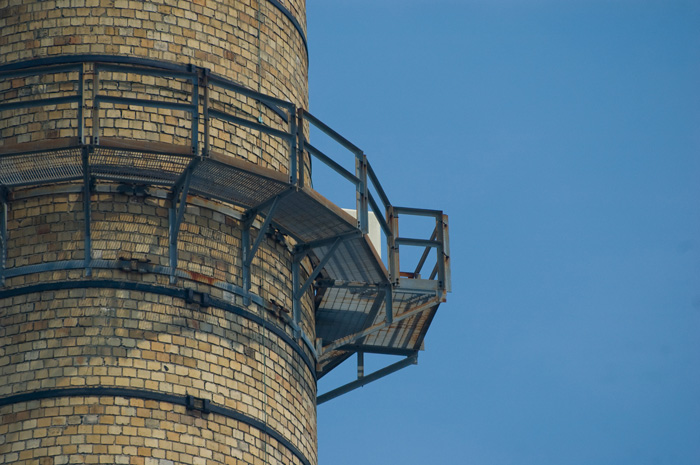 A yellow brick smokestack, with a service platform wrapped around it, stands out against a cloudless blue sky.